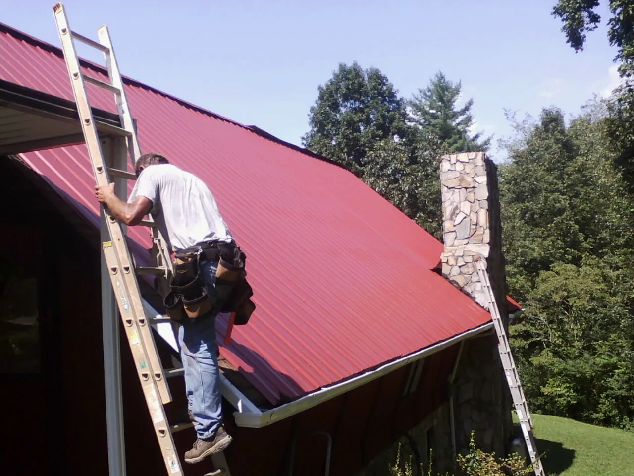 Roofer on a ladder beside a red metal roof and stone chimney — Soffit & Fascia Repair by Roofing Brothers in Cataldo